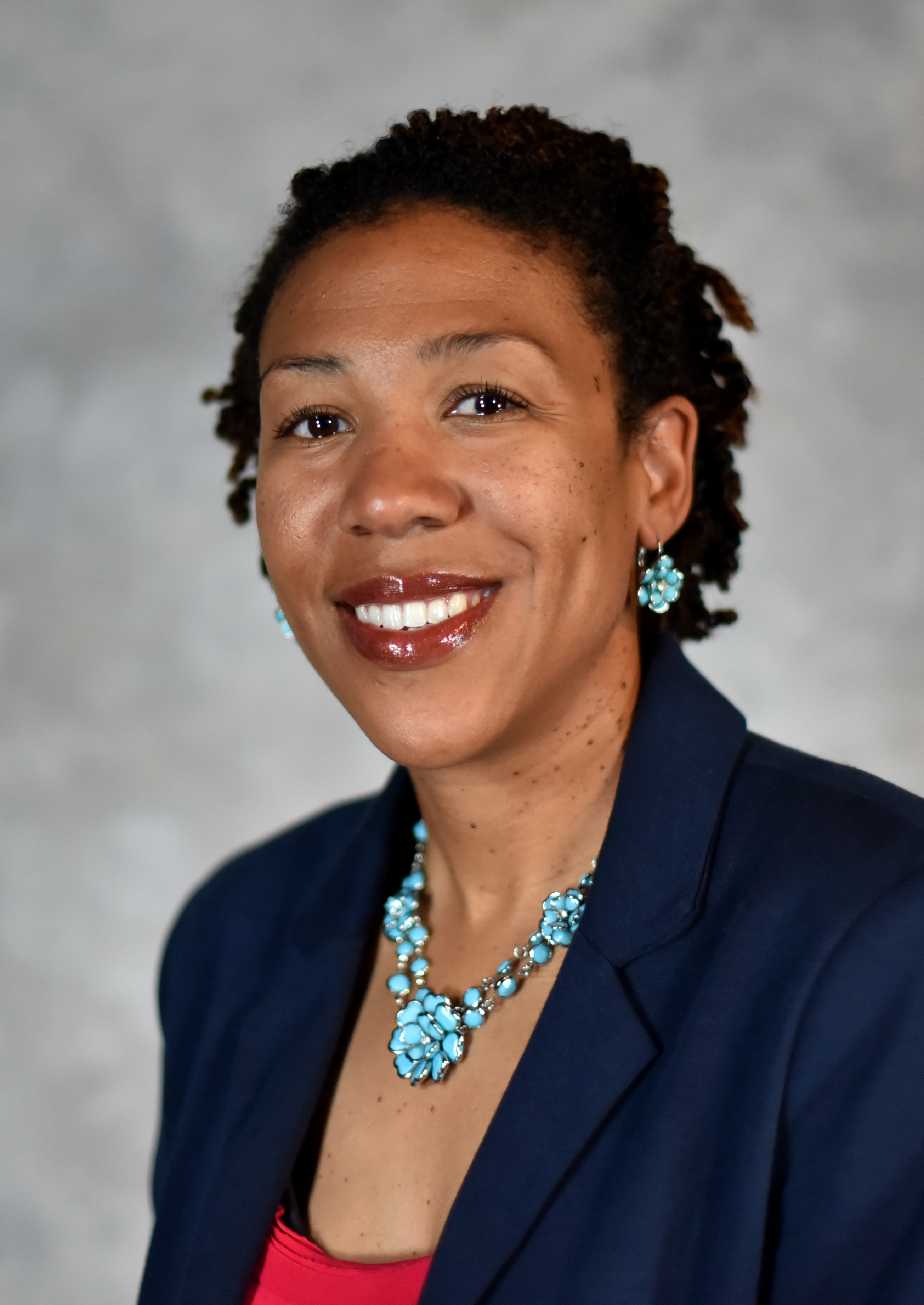 A headshot of Danica Sumpter, a black woman with dark brown, natural hair, smiling in a navy blazer. 